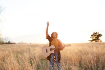 a woman standing in a field with a guitar with hands raised 