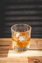 A glass of wiskey on the Rocks, on a black background, and a wooden table.