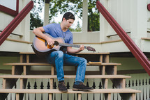 A young man plays a guitar outside in a gazebo rehearsing for worship music.