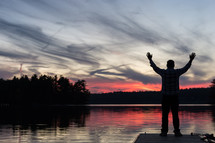 Silhouette of  a man with arms raised in praise at sunset over a lake.