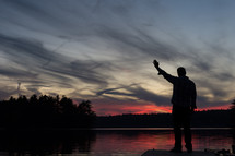 Silhouette of a man with arm raised in praise on a pier at sunset over a lake.