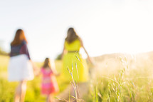 Mother and daughters holding hands while walking through a sunny field.