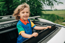 Adorable happy little boy stands in open car sunroof during road trip in countryside at summer. Concept of family leisure, active traveling. High quality photo