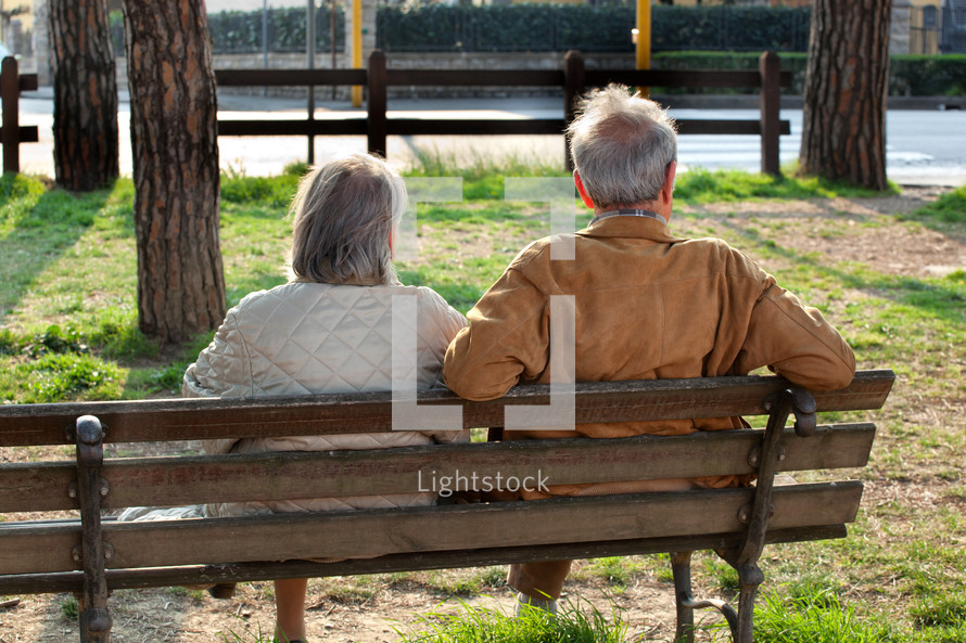 Elderly couple sitting on a bench on a sunny day at the park. They are from behind and are not recognisable.