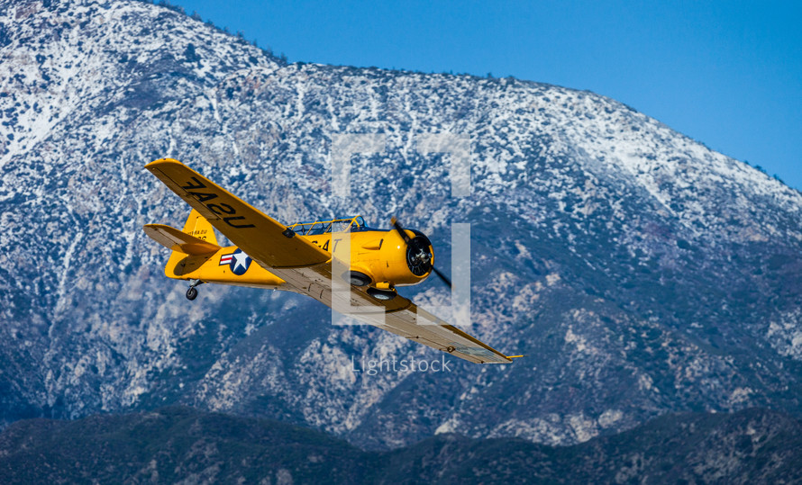 A yellow single engine airplane flying near a mountain.