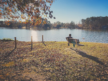 a boy sitting on a bench beside of a pond 