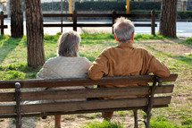 Elderly couple sitting on a bench on a sunny day at the park. They are from behind and are not recognisable.