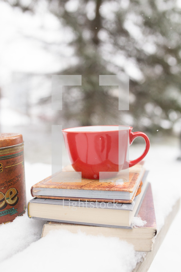red mug on a stack of books on a table outdoors in the snow 
