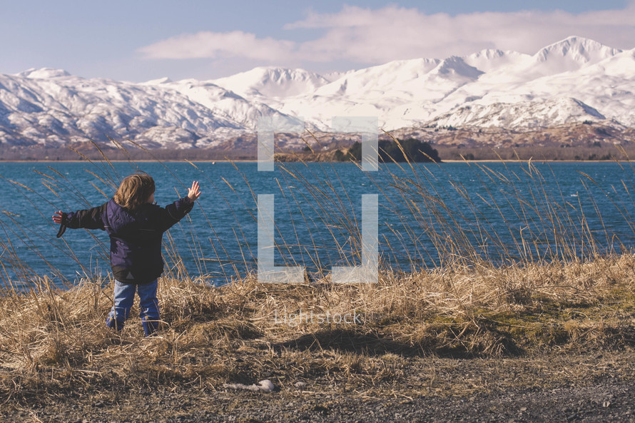 A child with arms outstretched stands before  a lake and a snow covered mountain range.