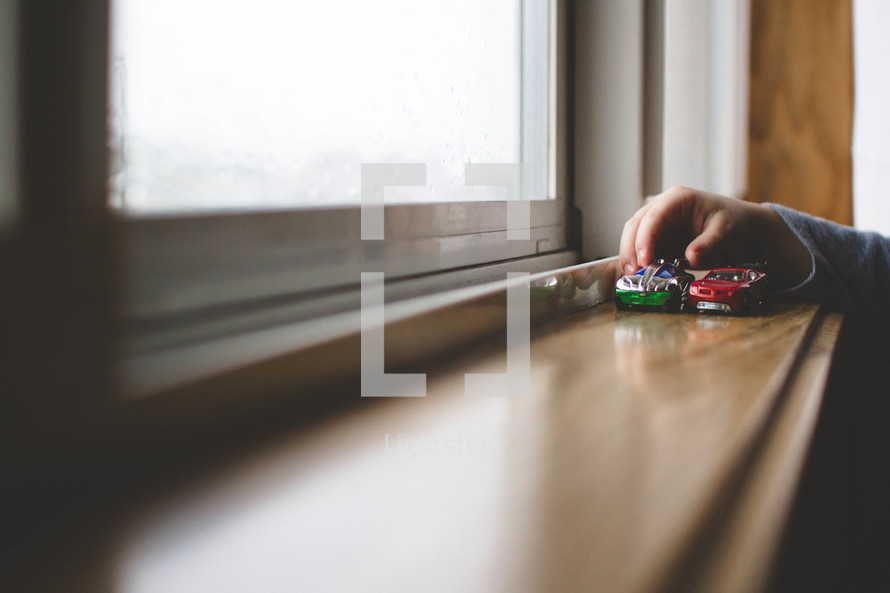 A child plays with toy cars on a windowsill.