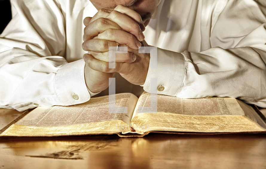 A married man in a white, long-sleeved shirt bows his head and has his hands clasped in deep prayer over his Holy Bible. (Model release on file)
