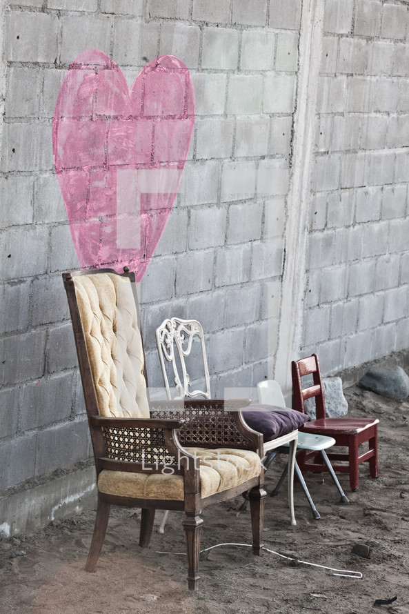 Pink heart painted on a stone wall of a building with several different kinds of chairs placed in front of the wall