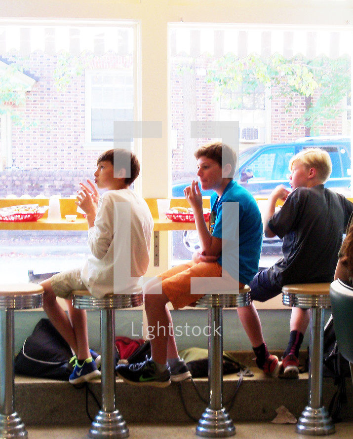 Soda Fountain Kids - A group of three young elementary school boys sit and relax together in a local soda fountain after school eating French fries, hamburgers and soda on a sunny day sitting on round stools in front of a large picture window in a local downtown soda shop restaurant. 