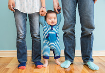 Infant child standing on the wood floor holding his parents' hands.