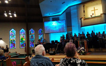 Church Members, audience and parishioners experience a multimedia show of sight and sound during a Sunday Morning Praise and worship service in church with blue animated screens and a full choir and worship band playing on stage and the Pastor baptizes someone from the church baptistry. 