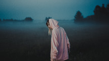 a young woman in a hoodie walking in a foggy field of tall grasses 