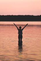 a female standing in lake water with arms raised 