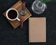A wristwatch and cup of coffee on a wooden board next to a spiral notebook and a plant in a vase.