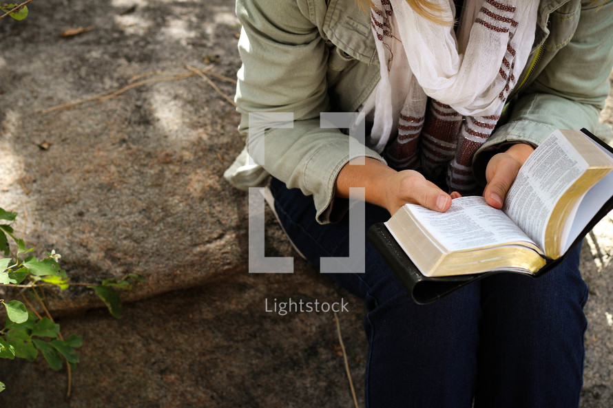 A woman sitting outdoors on a rock and reading a Bible.