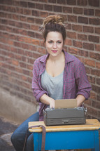 A young woman sitting at a school desk with a typewriter in front of a brick wall.