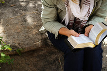 A woman sitting outdoors on a rock and reading a Bible.