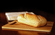 A rustic loaf of bread is on a wood cutting board in front of an open Holy Bible in the background.  Bread is powerful religous symbolism for Christianity, as a part of the Lord's Prayer asks the Father in heaven to "give us each day our daily bread" (Luke 11:3), along with Jesus Christ calling himself the "bread of life" in John 6:35.