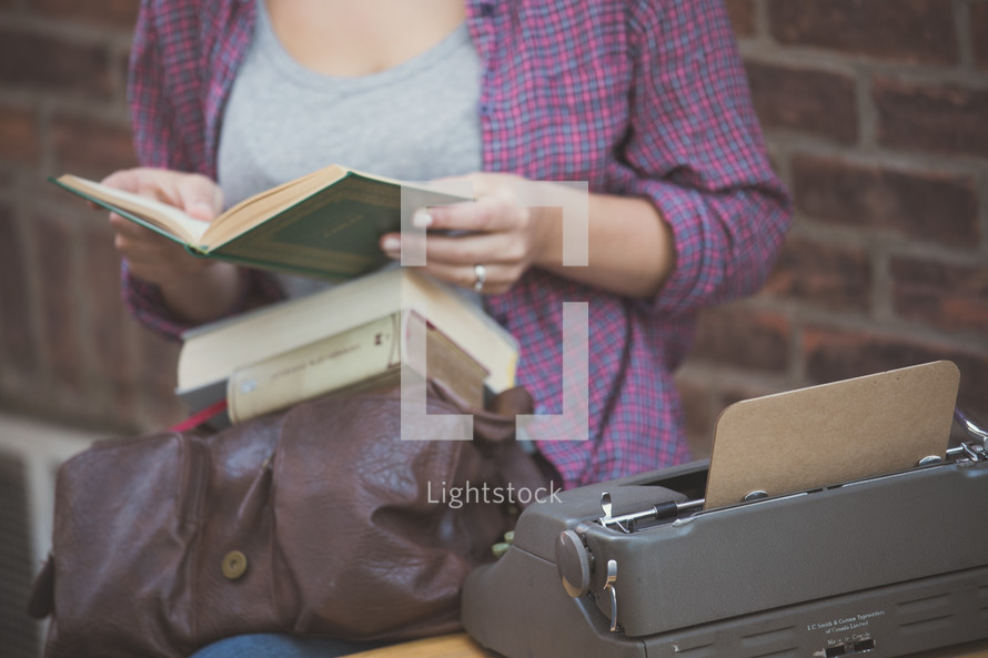 A young woman with a stack of books and a typewriter by a brick wall.
