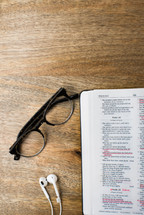 earbuds, reading glasses, and an open Bible on a wood table 