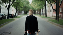 a young woman walking down a neighborhood street carrying a skateboard 