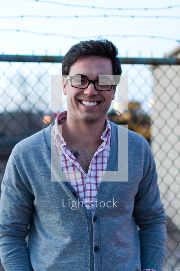smiling man in glasses standing in front of a chain linked fence