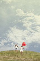 Girl running down a hill behind a girl holding red balloons.