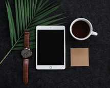 A sprig of green leaves, wristwatch, cell phone, cup of coffee and a blank notecard.