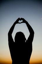 Silhouette of person making a heart shape with their hands with arms extened over head with sunset in background..