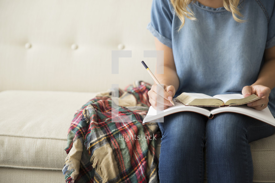 a woman reading a Bible and writing in a journal sitting on a couch 