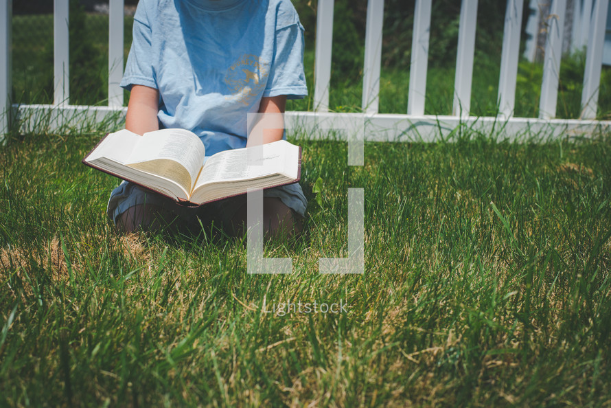 a boy kneeling in the grass reading a Bible 