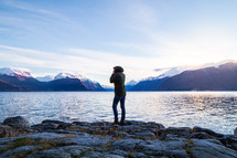 woman standing on rocks on a lake shore looking out at mountains
