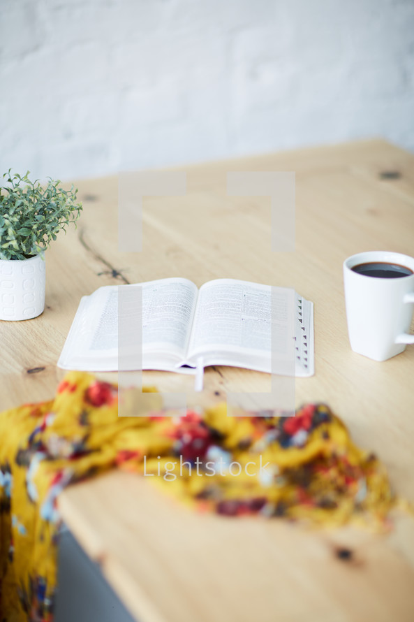 a Bible on a wood table, floral scarf, and coffee cup 
