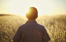 a young man walking through a field of tall grass at sunset 