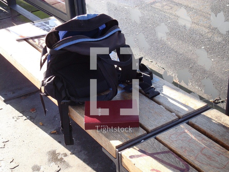 bible next to a backpack on a bench at a bus stop, 

bible, bench, stop, backpack, bus stop, waiting, tram, station, streetcar, trolley, transportation, way, bus, bag, rucksack, knapsack, booksack, daybag