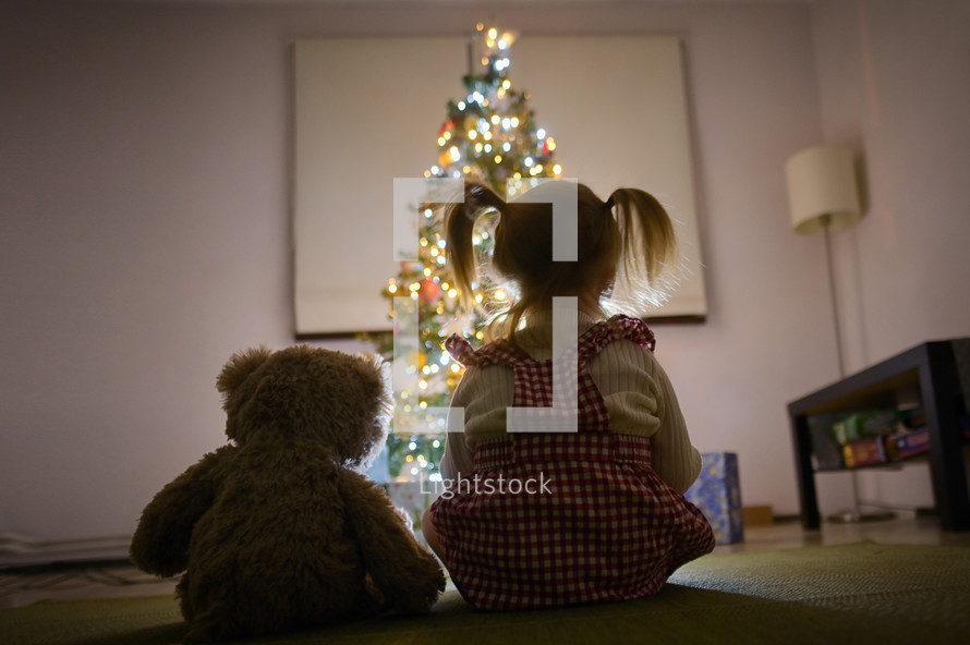 A Little Girl Looking At Christmas Tree With Her Teddy Bear At Home