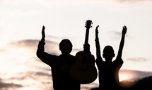 silhouettes of two boys at sunset one with a guitar and the other with hands raised 