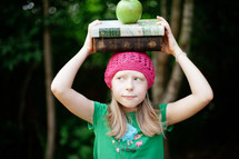 girl child holding a stack of books and an apple on her head