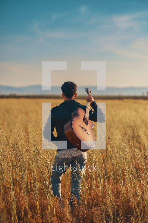 A young man standing in a field with a guitar over the shoulder