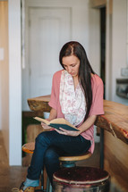 a young woman sitting on a stool in a kitchen reading a Bible 
