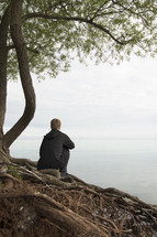 a man sitting on tree roots looking out at water 