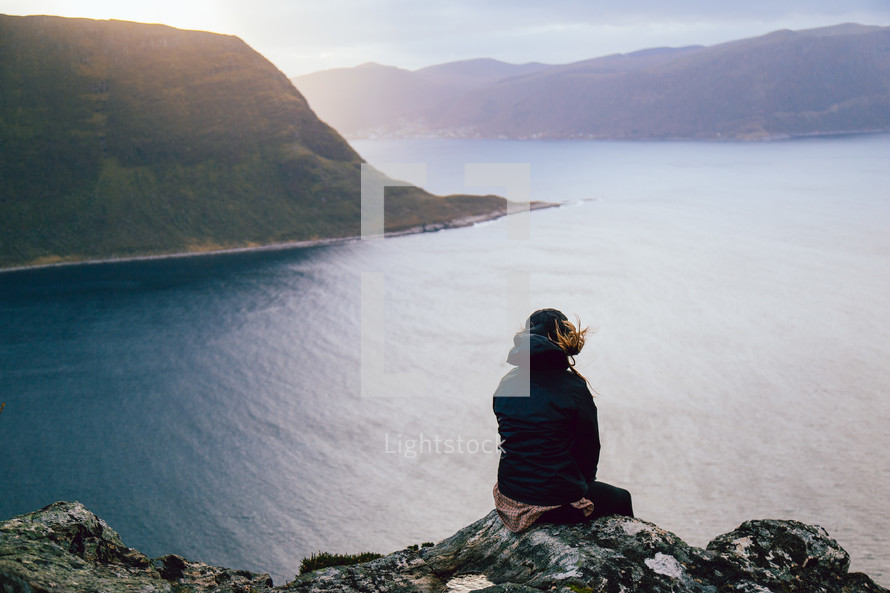 A person sitting on a high cliff overlooking a body of water surrounded by mountains.