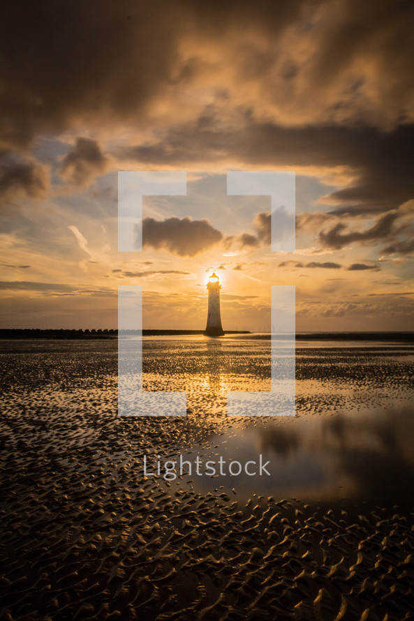Setting Sun through the lamp windows of a lighthouse on a beach at low tide.