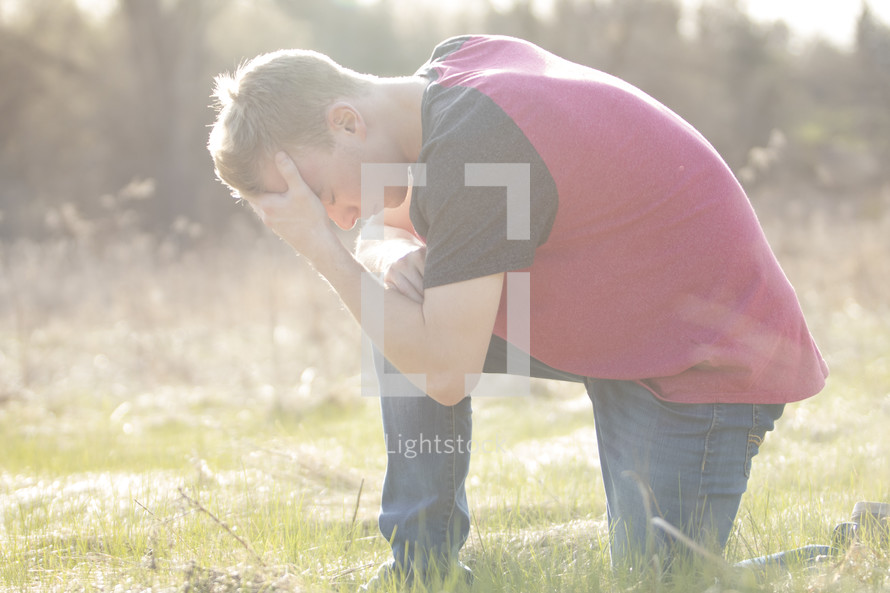 a man kneeling in grass with his hand on his forehead 