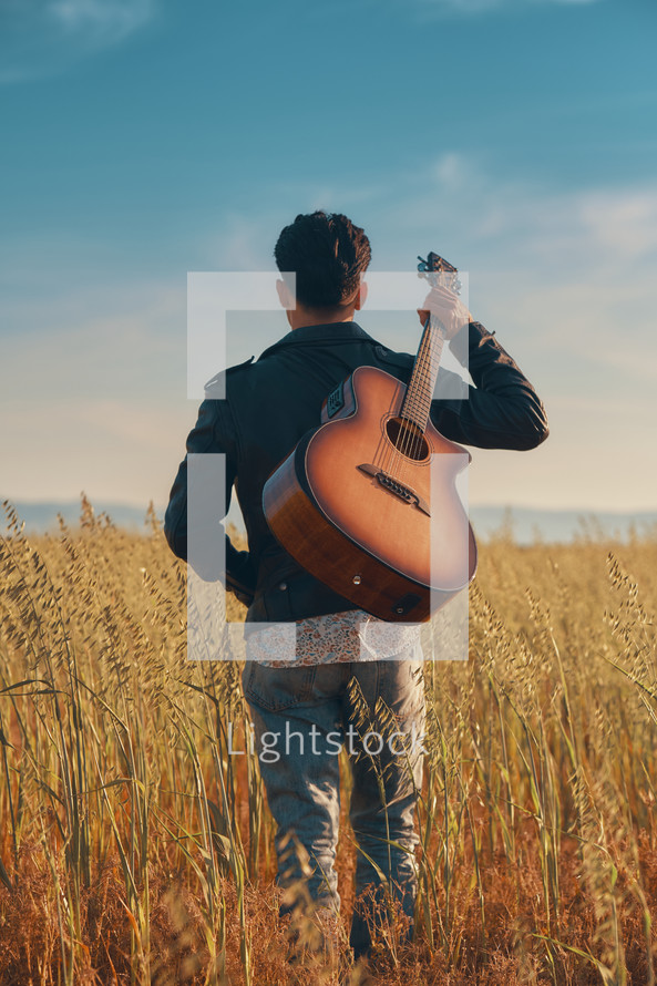 A young man standing in a field with a guitar over the back