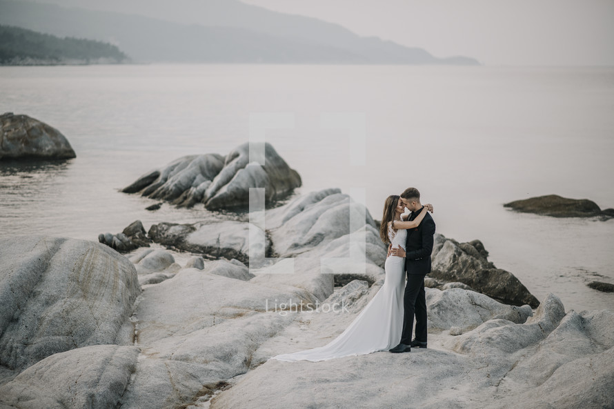 bride and groom standing on rocks along a shore 